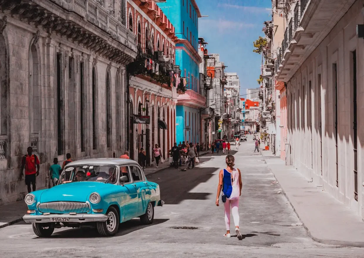 Woman walking down a colorful street in Havana, Cuba, with a vintage turquoise car and colonial buildings in the background.