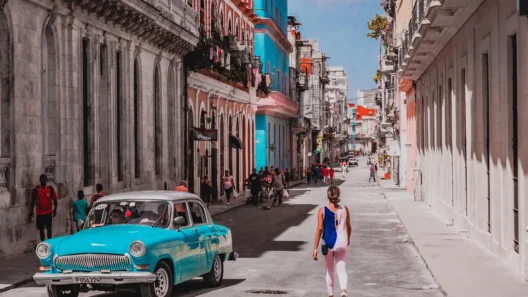 Woman walking down a colorful street in Havana, Cuba, with a vintage turquoise car and colonial buildings in the background.