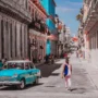 Woman walking down a colorful street in Havana, Cuba, with a vintage turquoise car and colonial buildings in the background.