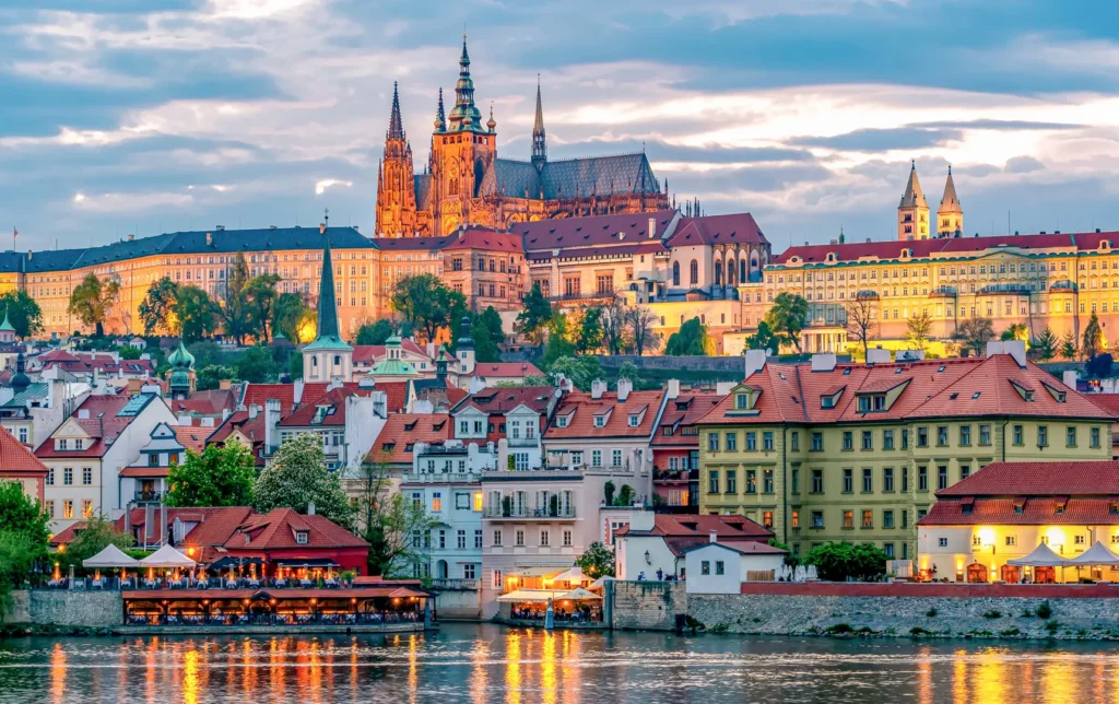 Prague Castle illuminated at sunset with colorful historical buildings and the Vltava River in the foreground.