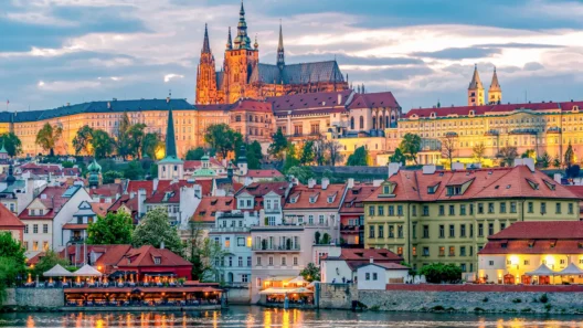 Prague Castle illuminated at sunset with colorful historical buildings and the Vltava River in the foreground.
