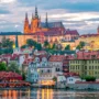 Prague Castle illuminated at sunset with colorful historical buildings and the Vltava River in the foreground.