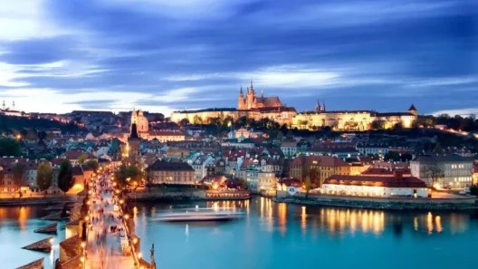 Evening view of Prague Castle and Charles Bridge illuminated over the Vltava River, with the city’s historic skyline and vibrant lights reflecting on the water.