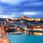 Evening view of Prague Castle and Charles Bridge illuminated over the Vltava River, with the city’s historic skyline and vibrant lights reflecting on the water.