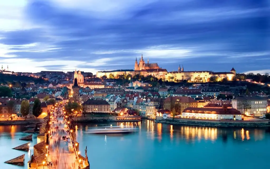 Evening view of Prague Castle and Charles Bridge illuminated over the Vltava River, with the city’s historic skyline and vibrant lights reflecting on the water.