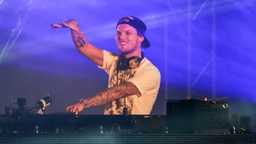 Avicii performing live on stage with headphones around his neck, smiling and raising his hand during a DJ set.