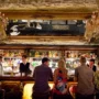 Elegant Prague cocktail bar with vintage chandeliers, mirrored ceiling, and bartenders in formal attire preparing drinks for guests at a lively, atmospheric bar counter.