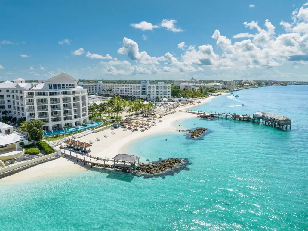 A stunning luxury beachfront resort in Nassau, Bahamas, with white sand, turquoise water, sun loungers, palm trees, and a wooden pier stretching into the crystal-clear sea under a bright blue sky.