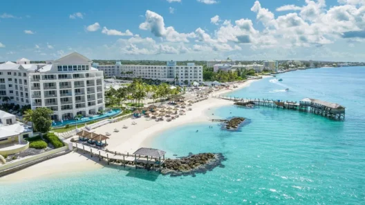 A stunning luxury beachfront resort in Nassau, Bahamas, with white sand, turquoise water, sun loungers, palm trees, and a wooden pier stretching into the crystal-clear sea under a bright blue sky.