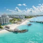 A stunning luxury beachfront resort in Nassau, Bahamas, with white sand, turquoise water, sun loungers, palm trees, and a wooden pier stretching into the crystal-clear sea under a bright blue sky.