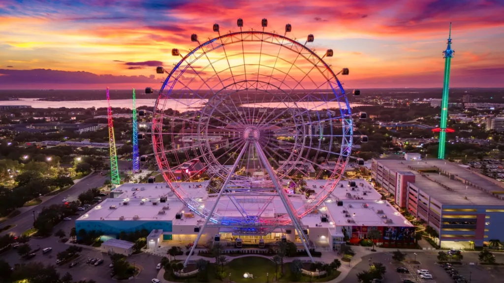 A colorful sunset view of The Wheel at ICON Park in Orlando, Florida, with its glowing lights and the city skyline stretching out toward the lake in the background.