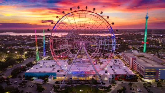 A colorful sunset view of The Wheel at ICON Park in Orlando, Florida, with its glowing lights and the city skyline stretching out toward the lake in the background.