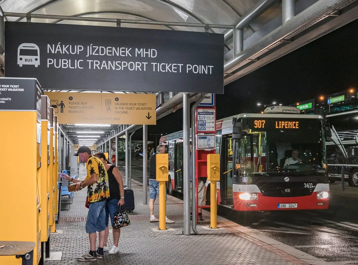 Travelers buying and validating public transport tickets at Prague Airport bus terminal at night.