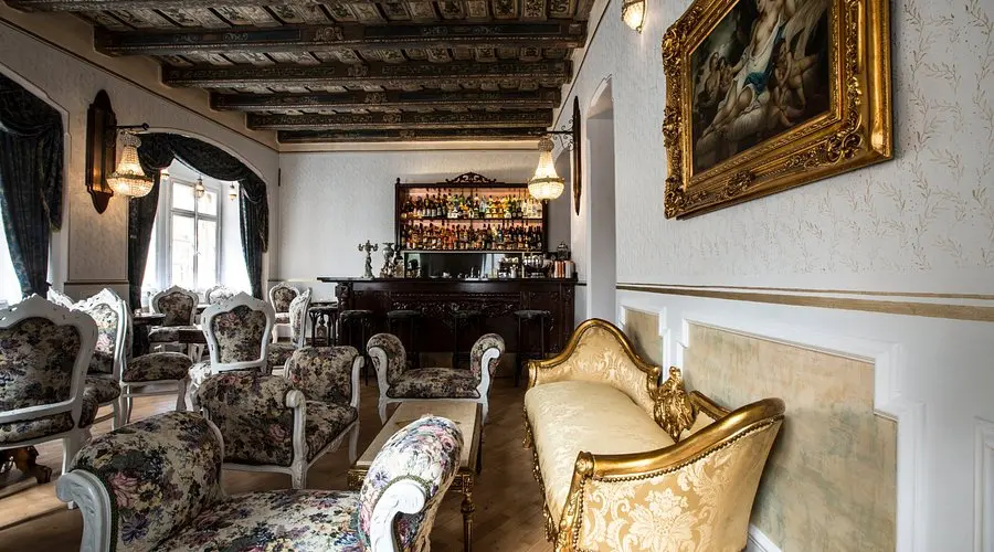 Luxurious Prague cocktail bar interior with ornate vintage armchairs, a golden sofa, classic chandeliers, and a decorative bar set beneath a painted ceiling and elegant wall art.