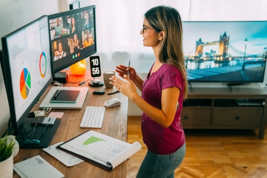 A woman working from her home office, leading a video call with colleagues on dual monitors, surrounded by charts, documents, and a London bridge photo on the TV in the background – a modern remote work setup.