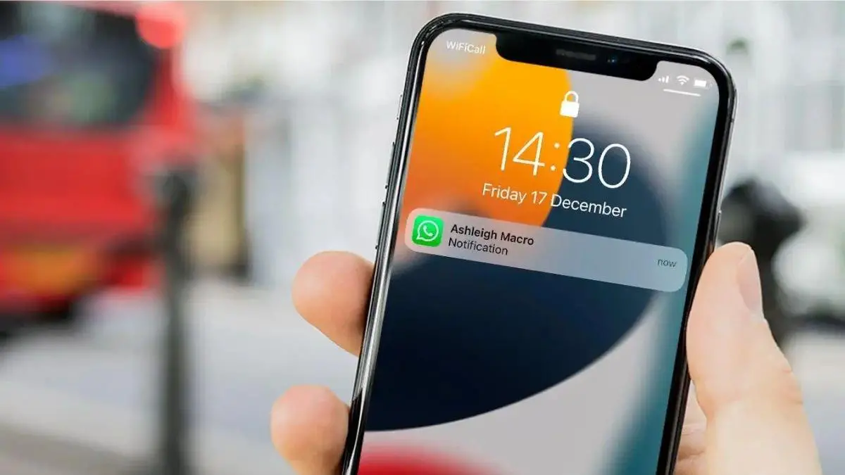 A hand holding an iPhone displaying a WhatsApp notification on the lock screen, with the time showing 14:30 and the background blurred.