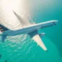 An American Airlines aircraft flying over the clear turquoise waters of the Bahamas. The sunlight reflects off the ocean surface, creating a tropical and serene atmosphere. The shot captures the plane from above, emphasizing the vibrant color of the sea below.