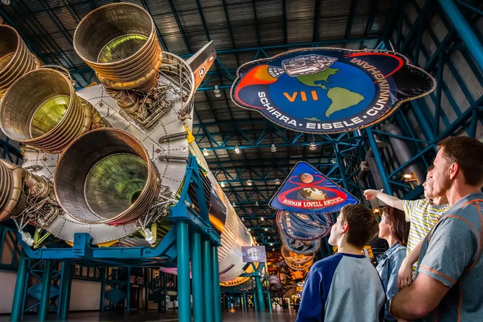 Visitors at Kennedy Space Center standing beneath the massive engines of a Saturn V rocket, looking up at colorful mission patches hanging from the ceiling in the Apollo/Saturn V Center.