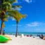 Sunny Florida beach with palm trees, colorful kayaks, and tourists sunbathing under a clear blue sky.