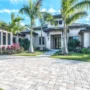 Modern tropical-style house with big windows, palm trees and a wide paved driveway under a bright blue sky.