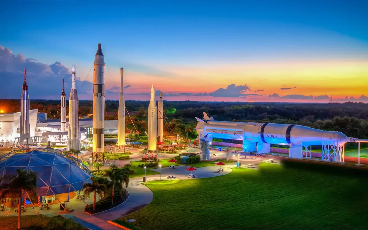 Colorful sunset view of the Rocket Garden at Kennedy Space Center, featuring towering historic rockets and Saturn IB under glowing lights, with palm trees and picnic tables in the foreground.