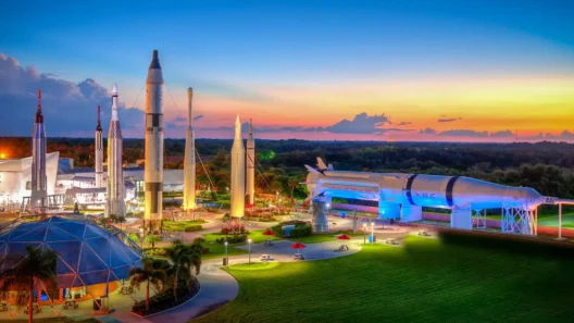Colorful sunset view of the Rocket Garden at Kennedy Space Center, featuring towering historic rockets and Saturn IB under glowing lights, with palm trees and picnic tables in the foreground.