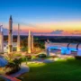 Colorful sunset view of the Rocket Garden at Kennedy Space Center, featuring towering historic rockets and Saturn IB under glowing lights, with palm trees and picnic tables in the foreground.