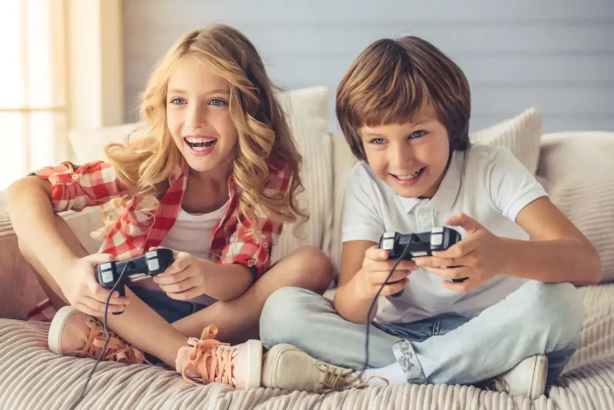 Two children sitting on a couch happily playing video games with wired controllers
