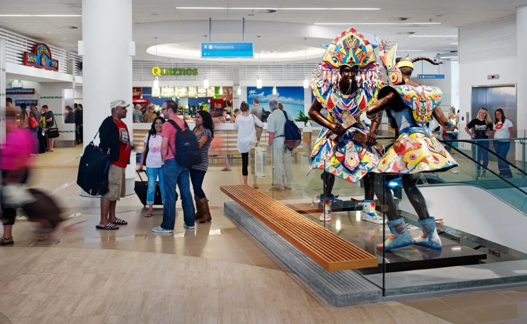 Lynden Pindling International Airport | Venclikovi.com The photo shows the lively interior of Lynden Pindling International Airport in Nassau, Bahamas. Travelers are walking through the terminal near a vibrant food court area featuring Quiznos, while two colorful Junkanoo statues stand proudly in the center, reflecting Bahamian culture. The scene captures the welcoming, artistic, and energetic vibe of the Bahamas from the moment you land.