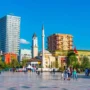 Skanderbeg Square in Tirana with the Clock Tower, Et’hem Bey Mosque, modern high-rises, and the statue of national hero Skanderbeg under a clear blue sky