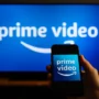 Hand holding a smartphone with the Amazon Prime Video logo in front of a large TV screen displaying the same interface, illustrating how VPN users stream Prime content across devices while traveling.