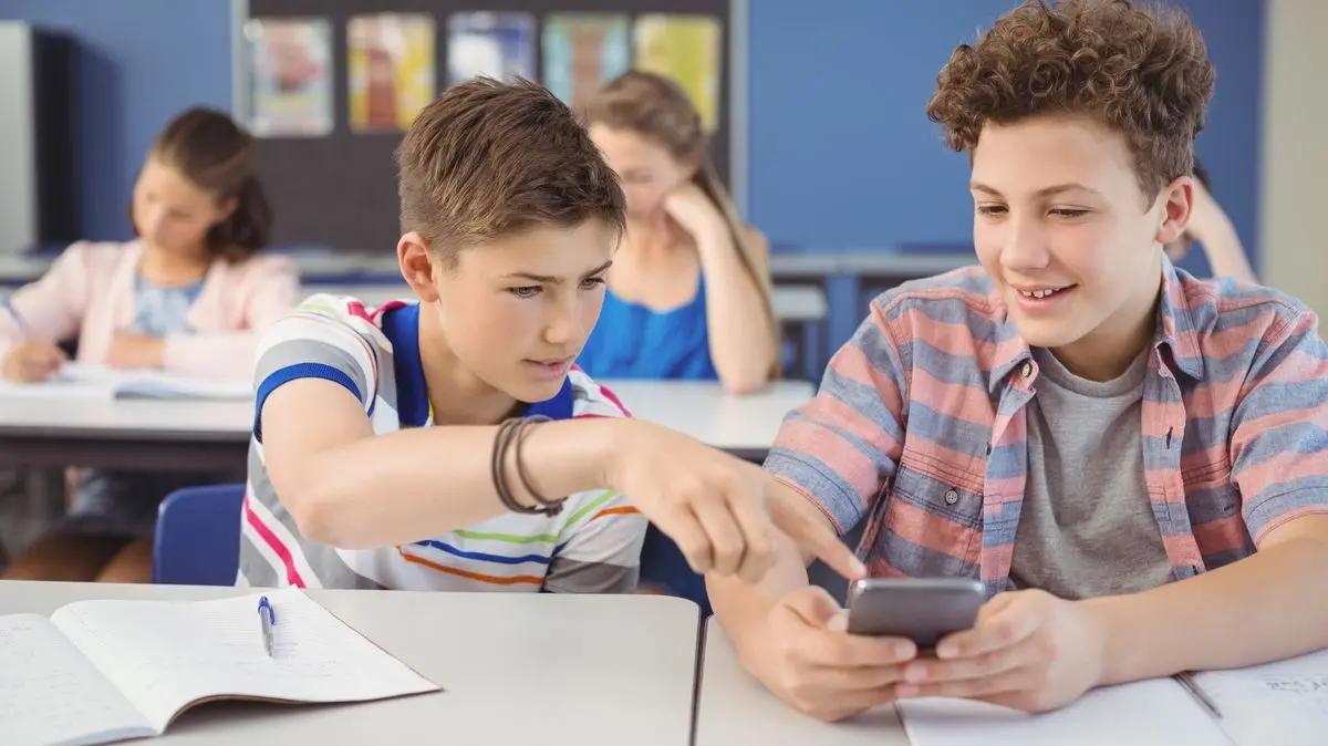 teens-phone-classroom-cyberbullying | Venclikovi.com Two teenage boys looking at a smartphone during class, sharing something privately while the rest of the students work, hinting at potential online teasing or gossip.