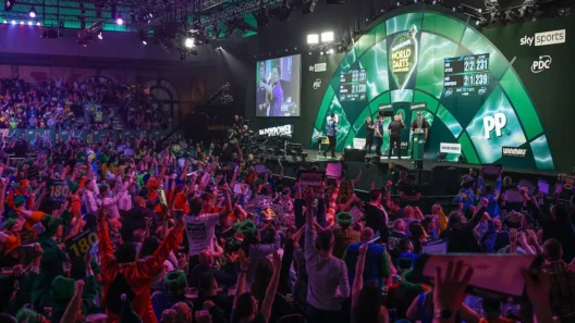 Crowd-packed Alexandra Palace during the 2025/26 World Darts Championship, with players on stage and fans celebrating a 180 moment during a live match streamed on Sky Sports and NOW TV.