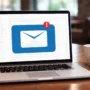 Laptop displaying an email inbox with a large envelope notification icon on the screen, placed on a table in a cafe setting, illustrating email access and alerts while working online.