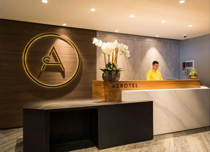 Aerotel London Heathrow | Venclikovi.com Modern minimalist reception desk of Aerotel London Heathrow at Terminal 3 Arrivals, featuring a warm staff greeting and clean, professional transit hotel branding