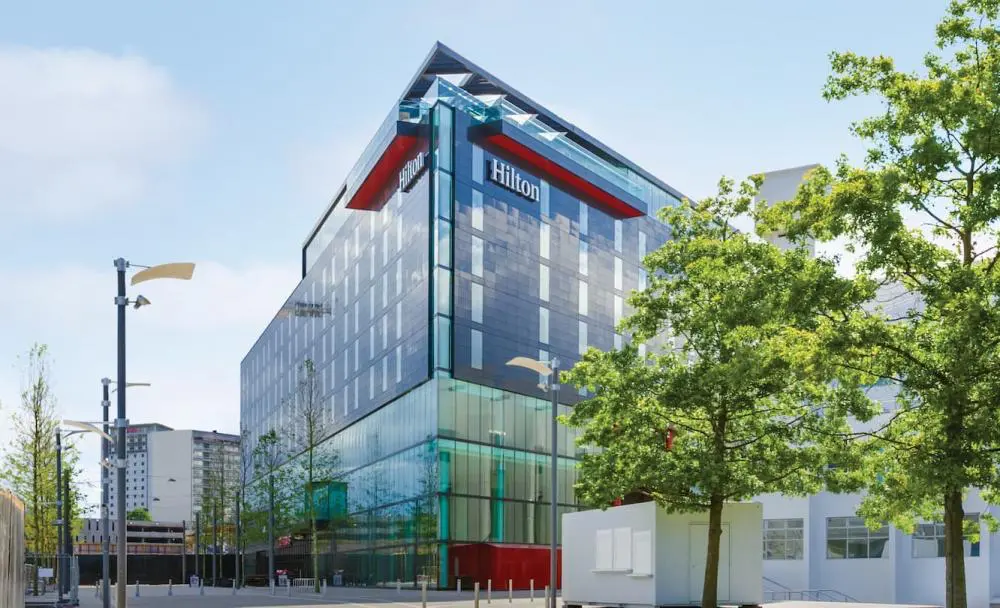 Hilton London Wembley | Venclikovi.com A modern multi-story Hilton hotel with a glass facade, situated next to green trees and a spacious pedestrian plaza near Wembley Stadium.