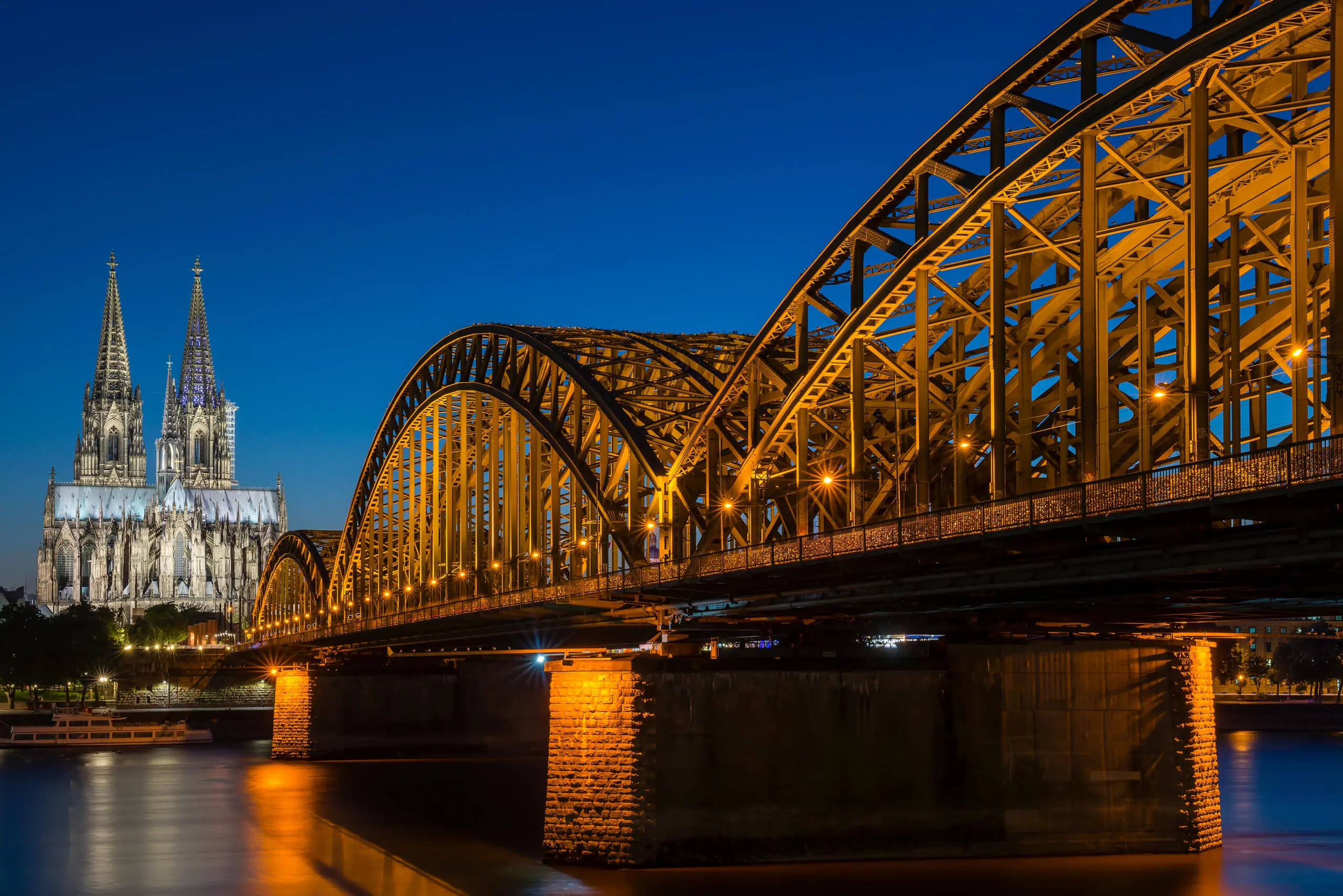 Hohenzollern Bridge | Venclikovi.com The Hohenzollern Bridge at night, brilliantly lit with golden lights, leading the eye toward the illuminated spires of the Cologne Cathedral.