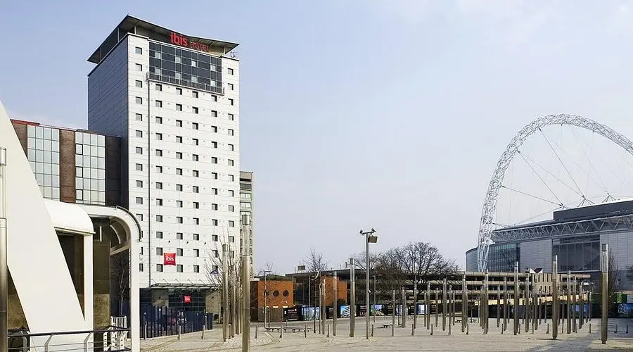 Ibis London Wembley | Venclikovi.com A tall, white high-rise hotel building with the red Ibis logo, with the iconic Wembley Stadium arch visible in the background.