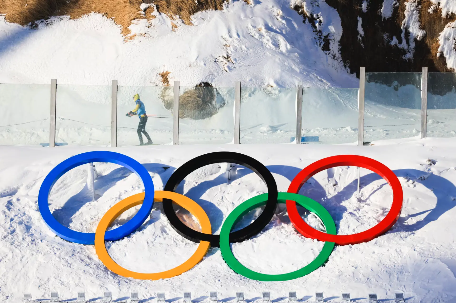 Olympic-Rings-Biathlon-2026-VPN-Streaming | Venclikovi.com Olympic Rings displayed in a snowy landscape during the Winter Games, with a cross-country skier in the background — symbolizing the 2026 Olympic biathlon atmosphere.