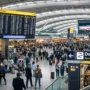 Busy interior of London Heathrow Terminal 5 departures area with travelers walking under a curved glass ceiling and large flight information boards.
