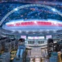 A stunning night-time aerial view of Wembley Stadium with its massive arch illuminated in white light and the bowl glowing red.