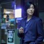 A tense nighttime scene from As You Stood By showing a man in a dark suit standing outside a small Korean shop, holding a lighter and looking off to the side with a worried expression.